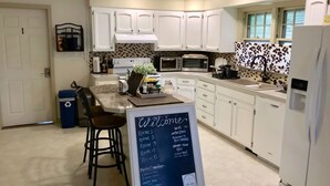 Une cuisine moderne avec des meubles blancs, un îlot central et un tableau noir indiquant les équipements disponibles, comme le Wi-Fi et le petit-déjeuner.