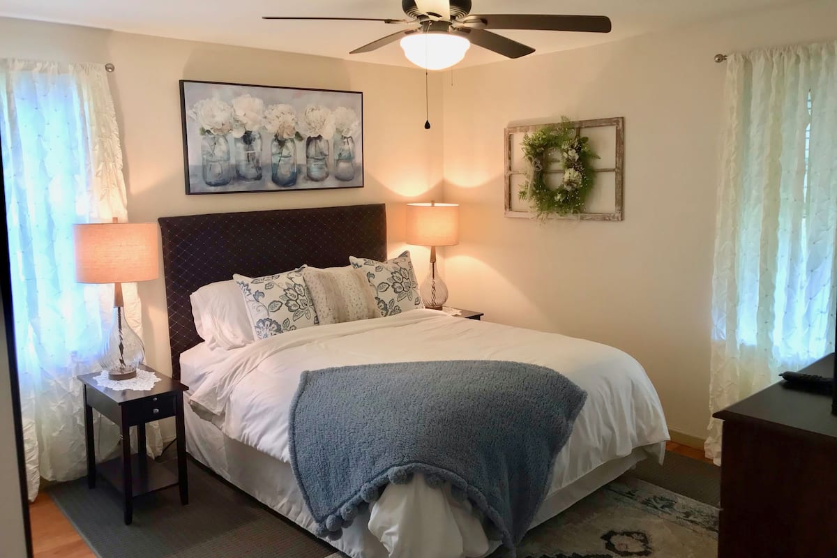 A bedroom with a bed, two bedside lamps, a ceiling fan, a painting, and a wreath on the wall.
