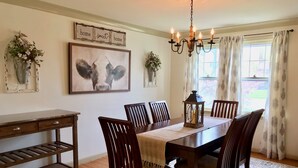 Une salle à manger avec une table en bois, six chaises, un lustre, un tableau encadré et une fenêtre avec des rideaux.