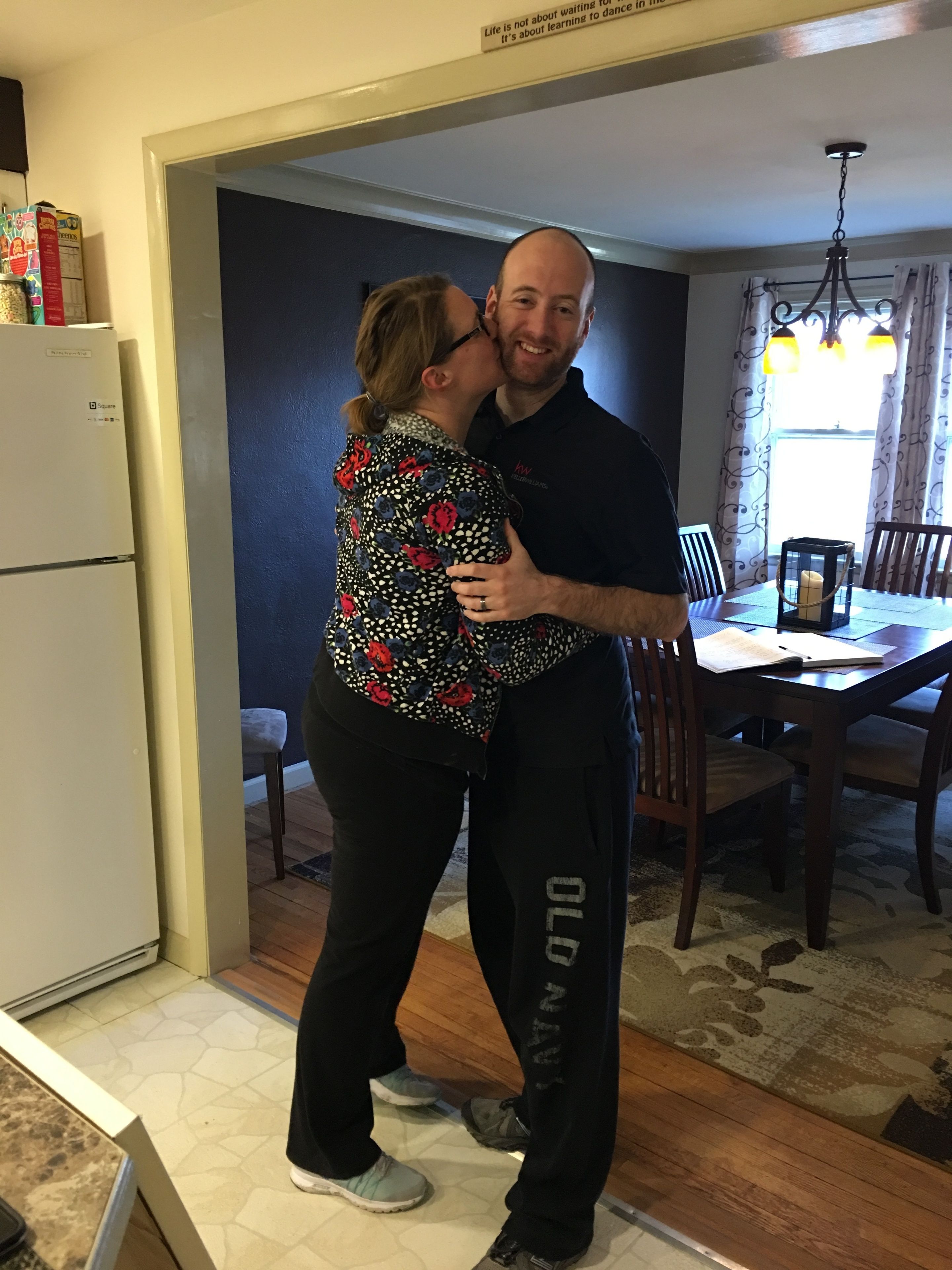 A couple embracing in a kitchen with a refrigerator, dining table, and chandelier.