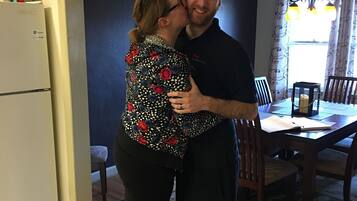 A couple embracing in a kitchen with a refrigerator, dining table, and chandelier.