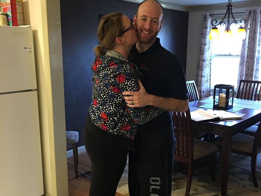 A couple embracing in a kitchen with a refrigerator, dining table, and chandelier.