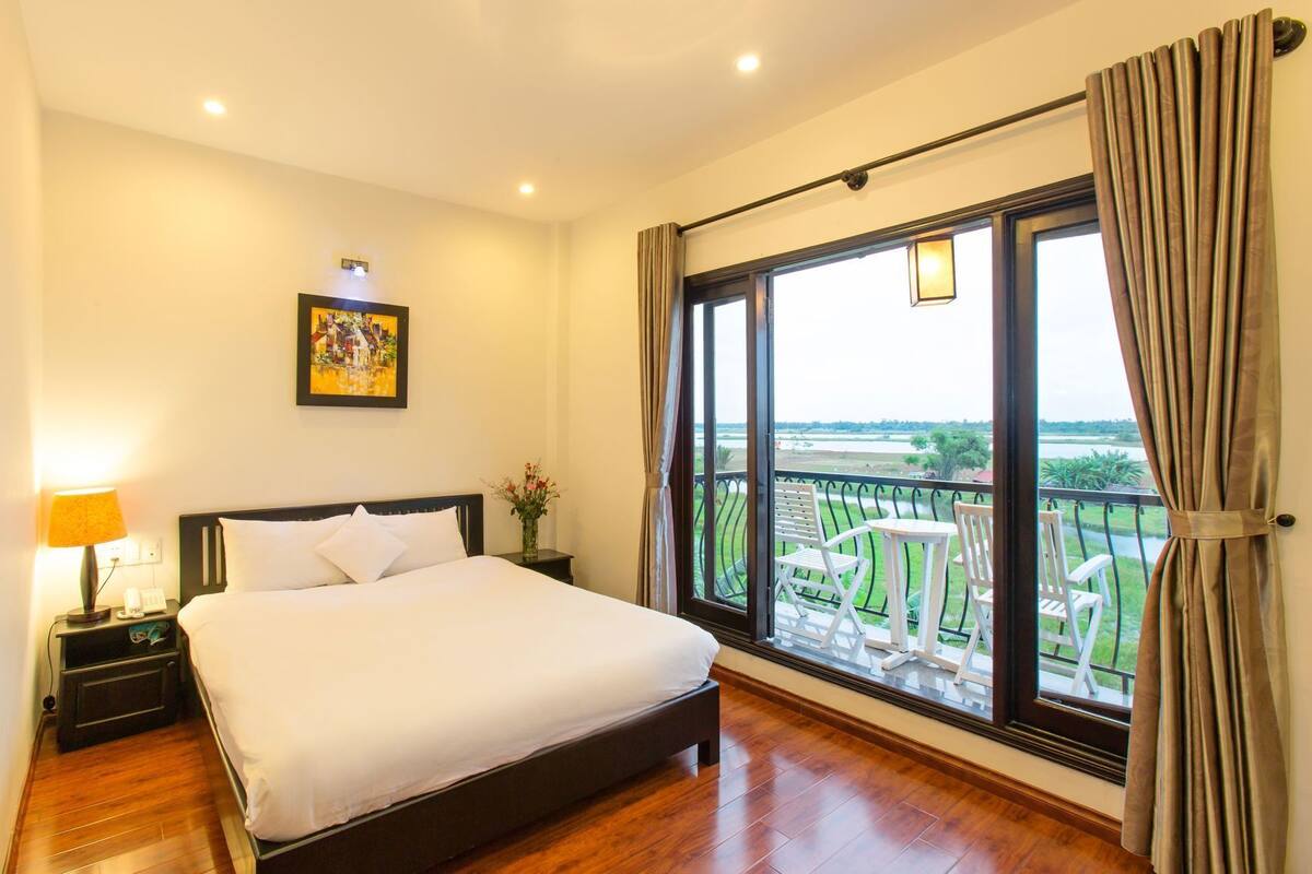 A hotel room with a large bed, a nightstand, a lamp, a painting, and a balcony with outdoor furniture.