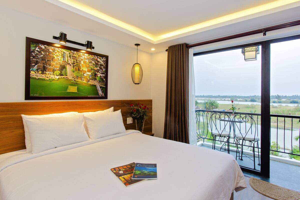 A hotel room with a large bed, a framed picture on the wall, a balcony with chairs, and a view of a river.