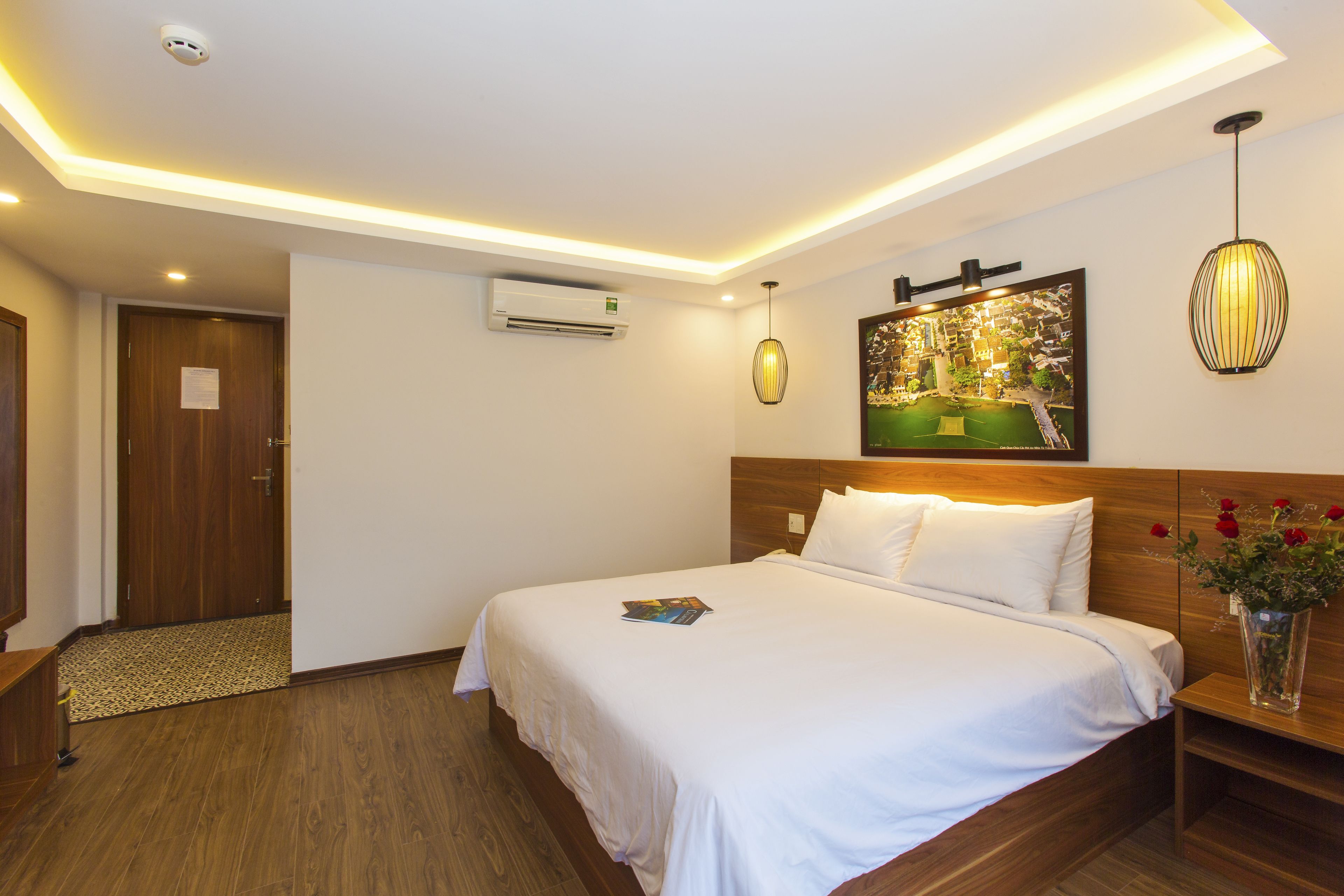 A hotel room with a large bed, a wooden headboard, a nightstand, a wall-mounted air conditioner, a framed picture, and a hanging pendant light.