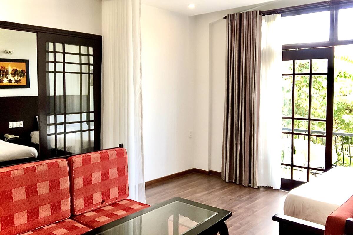 A hotel room with a bed, a glass-top coffee table, a red and white checkered sofa, a painting on the wall, and a view of greenery through the window.