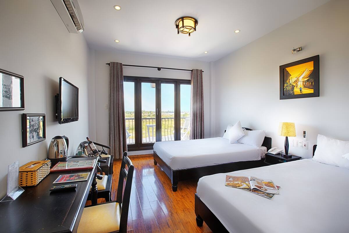 A hotel room with two beds, a desk with a chair, a television, and a large window with curtains.