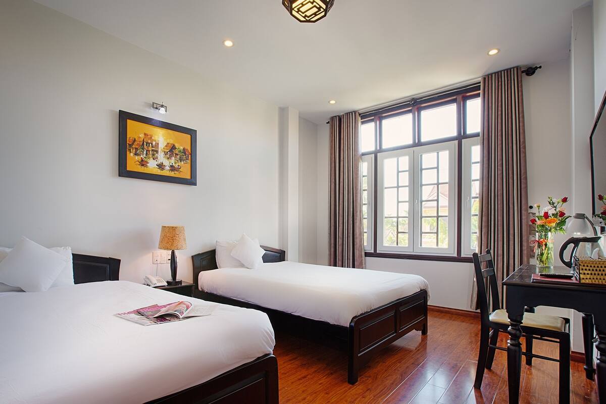 A hotel room with two beds, a desk with a lamp, a chair, a window with curtains, and a painting on the wall.