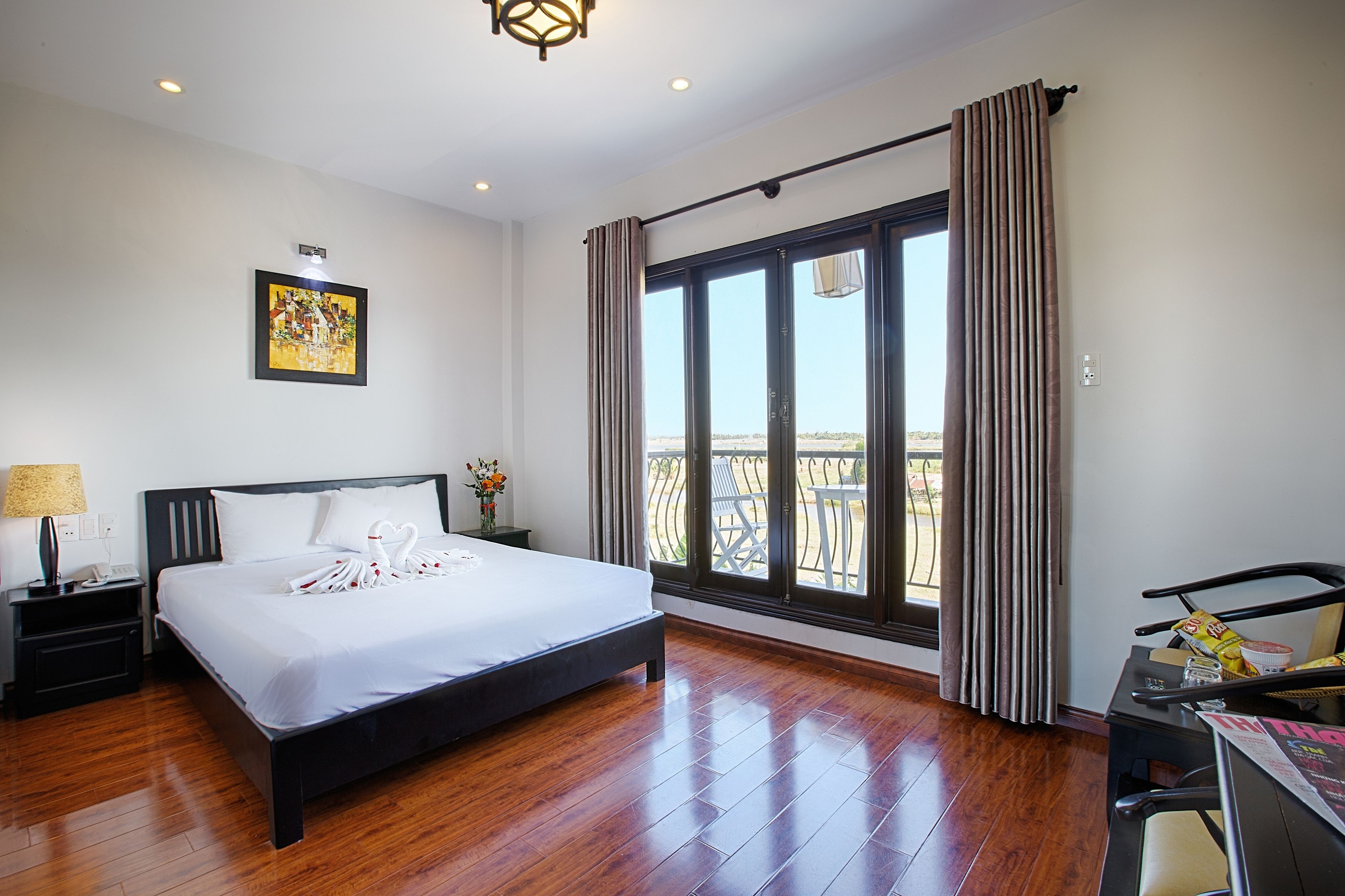 A hotel room with a large bed, a nightstand, a desk with a chair, a window with curtains, and a view of the outdoors.