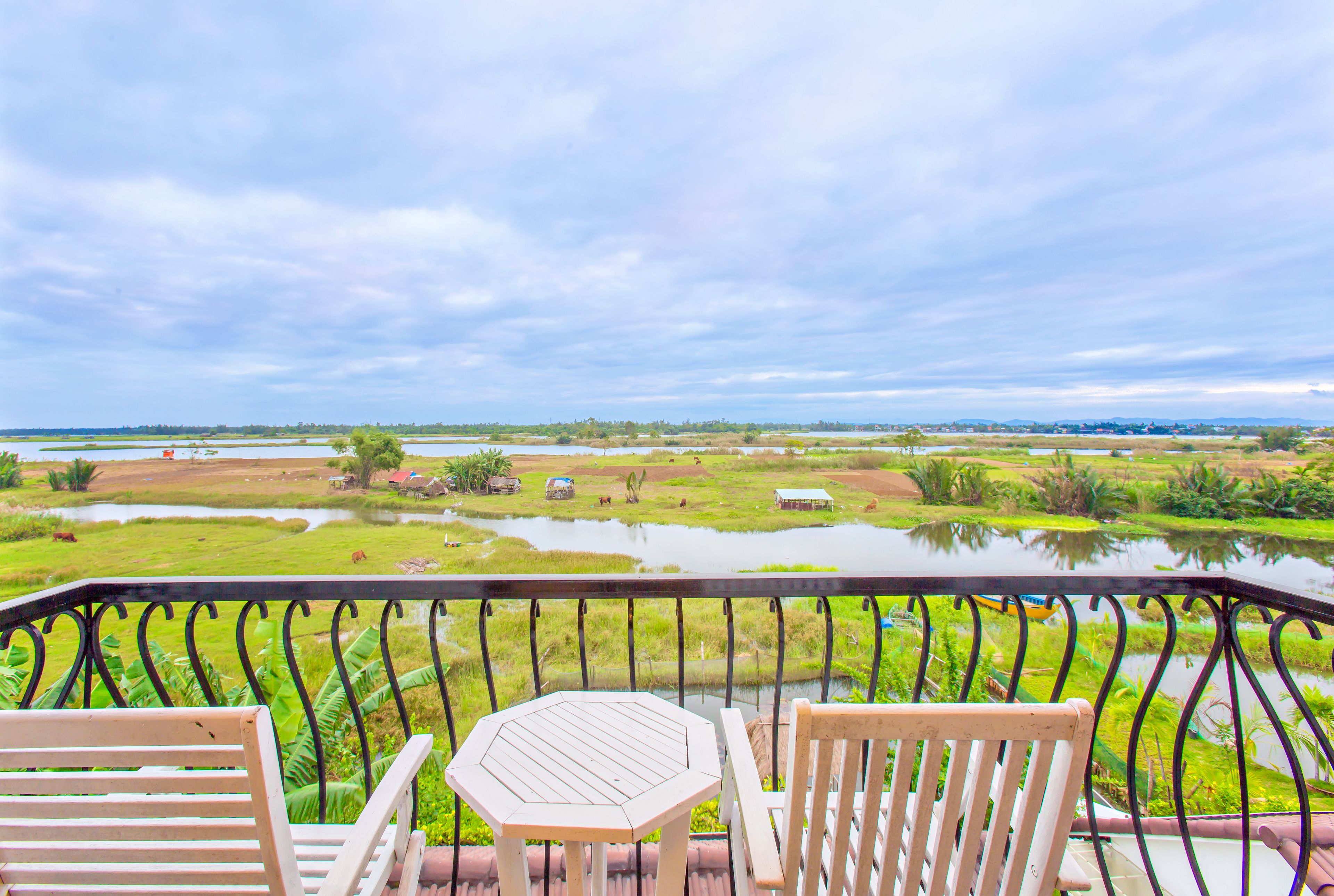 A balcony with two wooden chairs and a small table, overlooking a rural landscape with fields and a water body.
