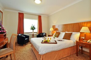 Une chambre d’hôtel avec un grand lit, deux tables de chevet avec des lampes, une chaise, une petite table avec un vase de fleurs et une fenêtre avec des rideaux.