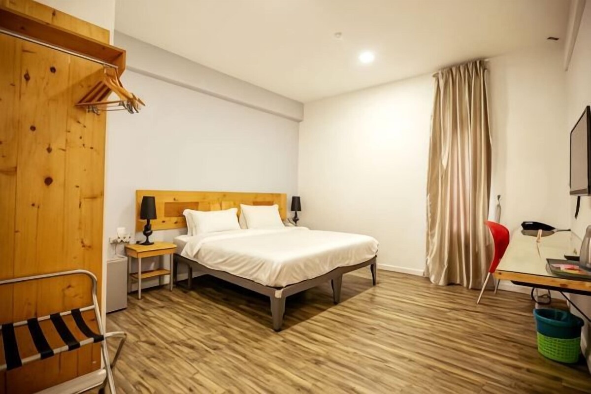 Een hotelkamer met een houten vloer, een bed met witte lakens, een nachtkastje met een lamp, een bureau met een stoel, en een rode stoel.
