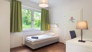 A small, neatly arranged bedroom with a single bed, a desk, and a chair. There is a window with curtains, a lamp on the desk, and a door with a sign.