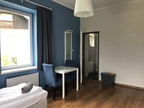 Ein modernes Schlafzimmer mit Bett, Schreibtisch und Stuhl. Ein Fenster mit Vorhängen, eine Tür zu einem Badezimmer und ein kleiner Mülleimer sind ebenfalls vorhanden.