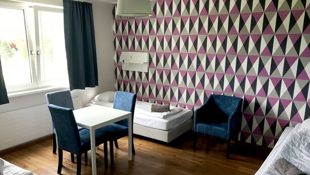 A room with a wooden floor, a white table with blue chairs, a bed with white bedding, and a patterned wall.