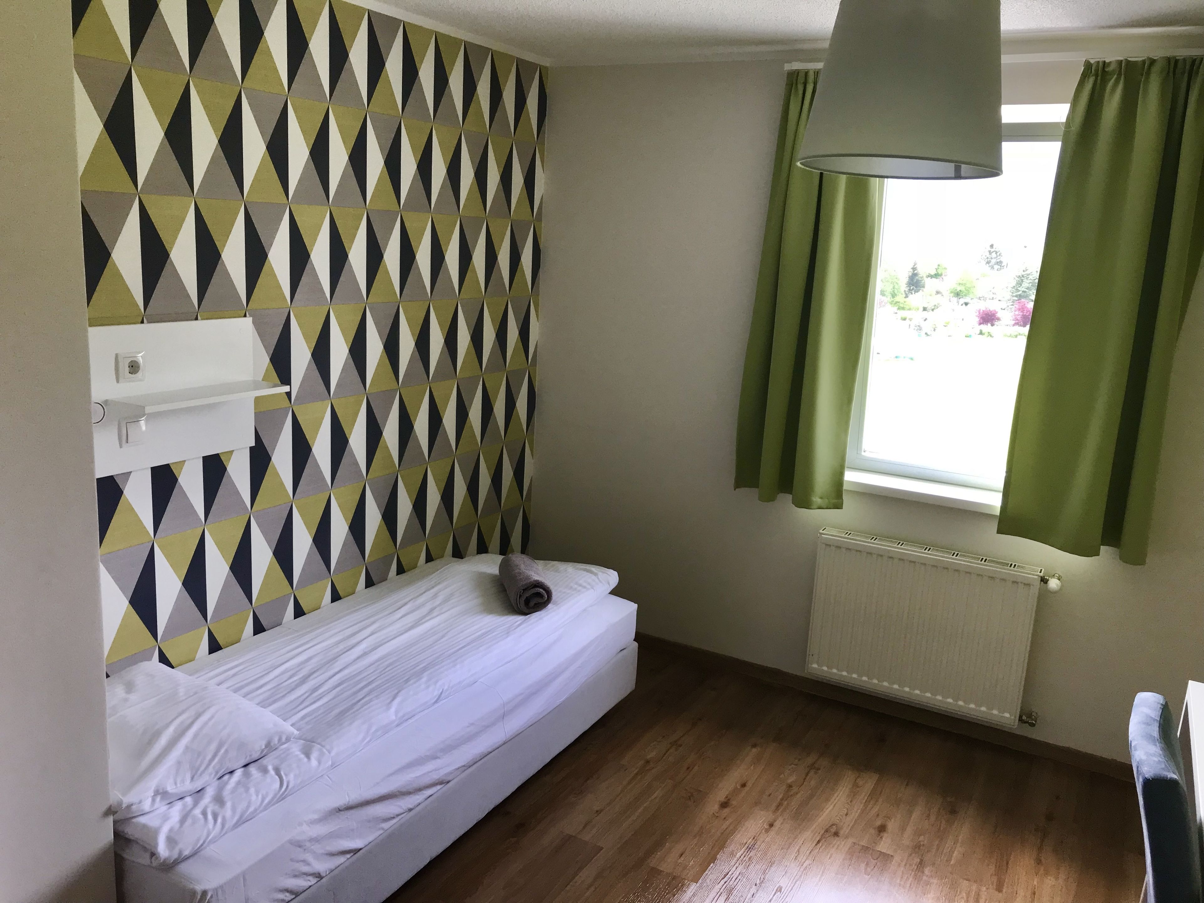 Une petite chambre avec un lit simple, un mur orné d’un motif géométrique, une fenêtre avec des rideaux verts et un radiateur.