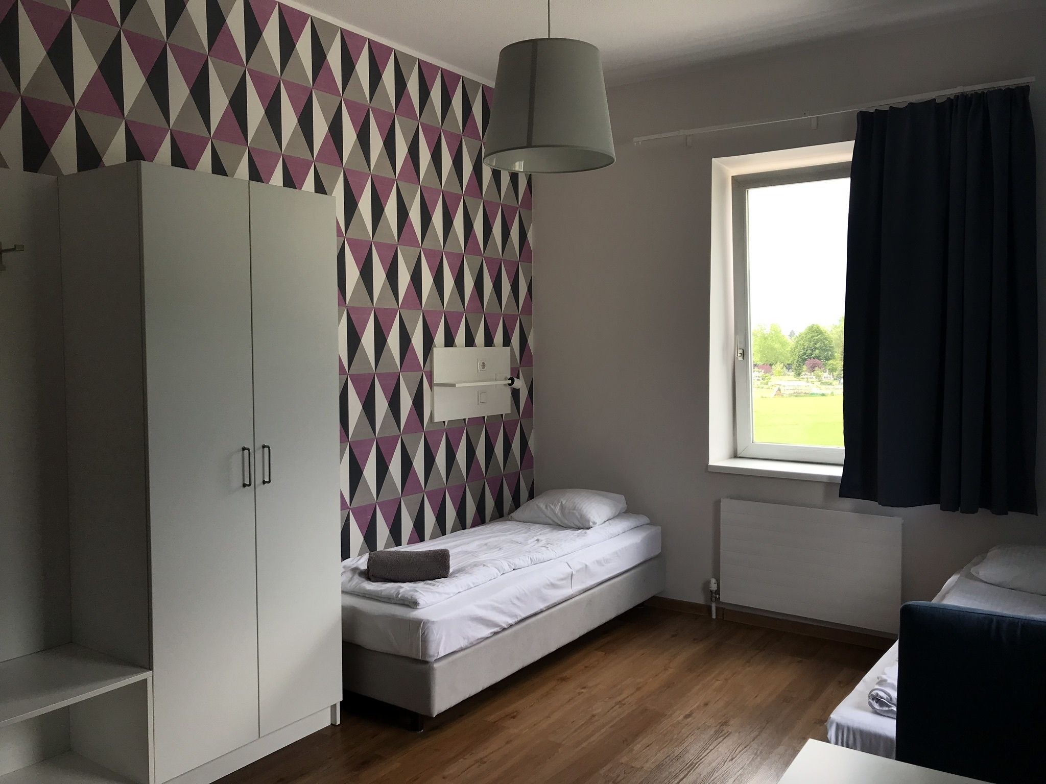 A bedroom with a bed, a wardrobe, a window with curtains, and a geometric-patterned wall.