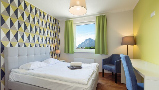 A hotel room with a bed, a desk, a chair, a window with curtains, and a geometric-patterned wall. - Marmota Hostel (Innsbruck)