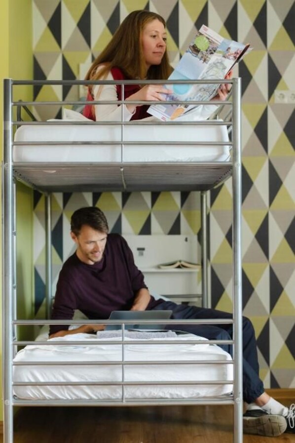 Una cama litera con dos niveles, una persona leyendo una revista en la cama superior y otra usando una computadora portátil en la inferior.