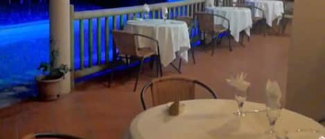 An outdoor dining area with round tables and chairs, covered by a roof with arches, and a stone wall in the background.