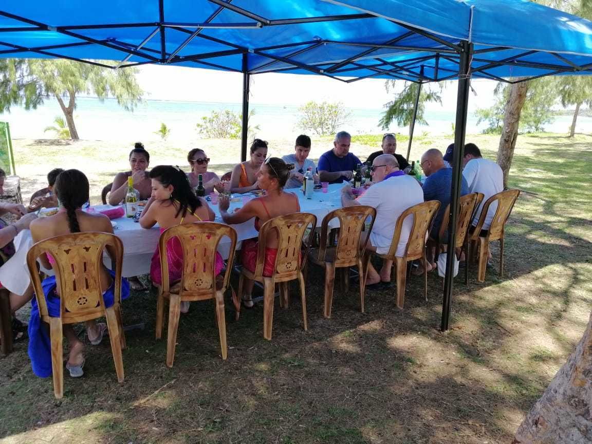 A group of people dining under a blue canopy at a beachside restaurant with wooden chairs and a white tablecloth.