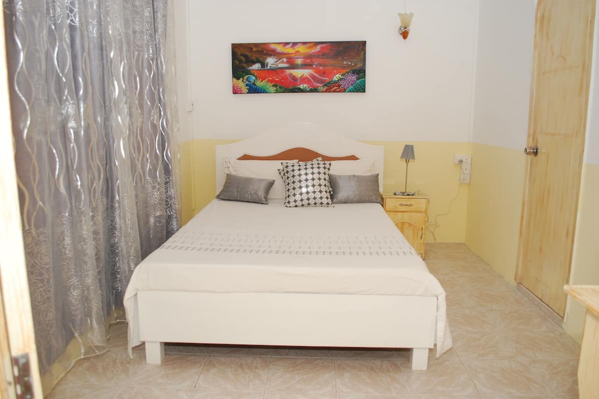 A bedroom with a bed, a nightstand, a lamp, a painting, and a wooden door.