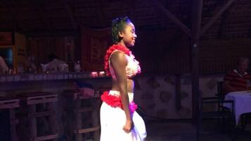 A person in traditional attire performing on a stage with a patterned floor, wooden ceiling beams, and tables in the background.