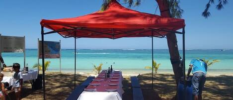 A beachside dining setup with a red canopy, tables set for two, and a view of the ocean.