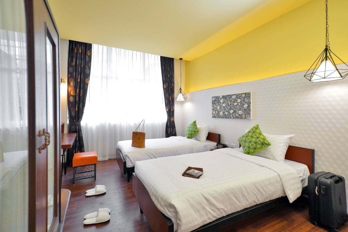 A hotel room with two beds, a wooden bench, a suitcase, and a window with curtains.