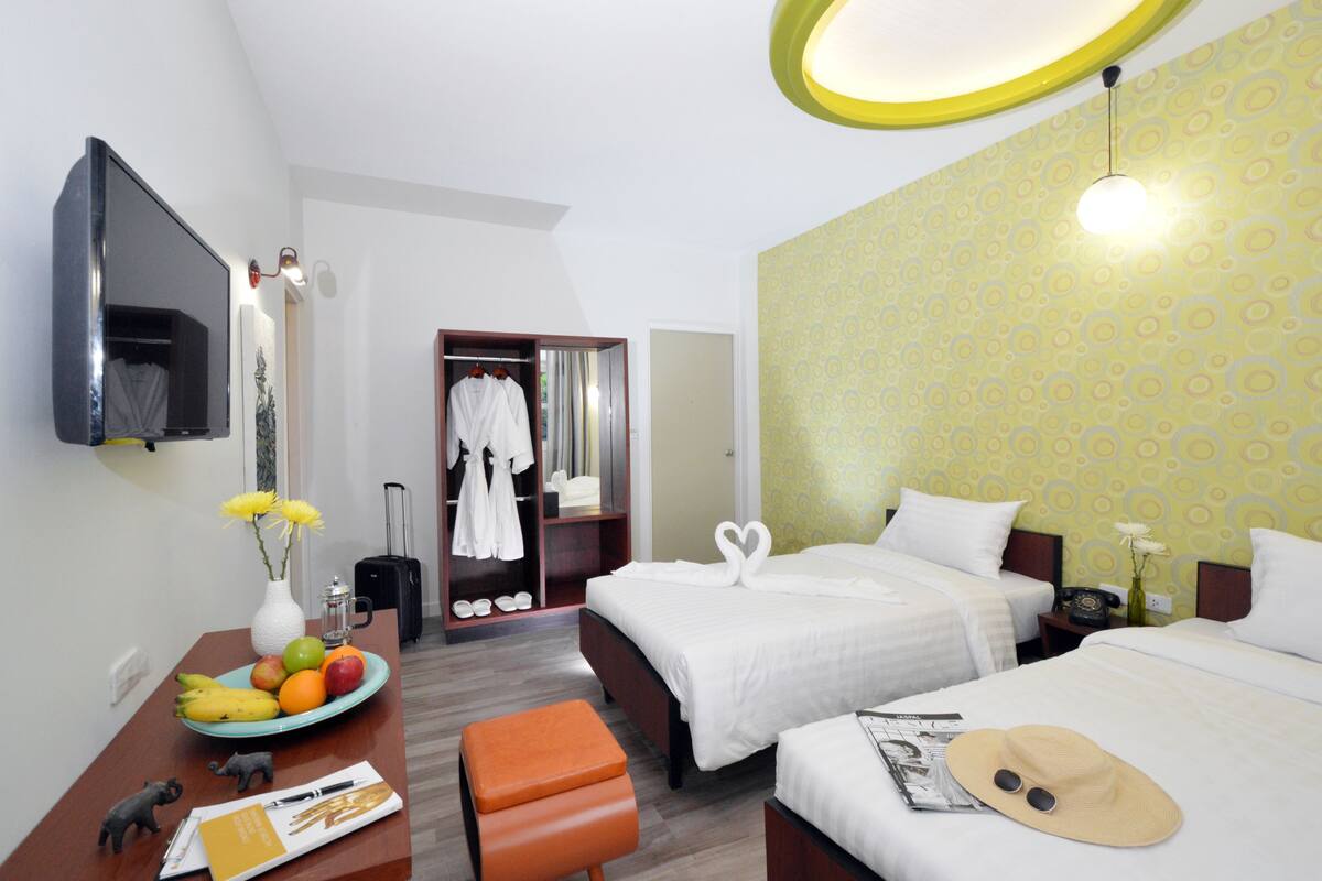 A hotel room with two beds, a desk with a fruit bowl, a suitcase, and a TV on the wall.
