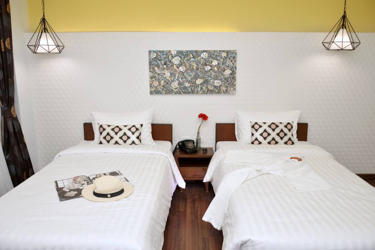 A hotel room with two beds, each with a patterned pillow and a white bedspread. There is a painting on the wall above the beds and a vase with a red flower on the nightstand between the beds.
