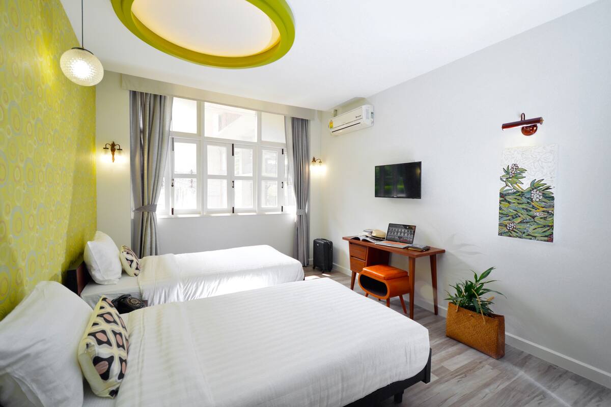A hotel room with two beds, a desk, a chair, a television, an air conditioning unit, and a window with curtains.