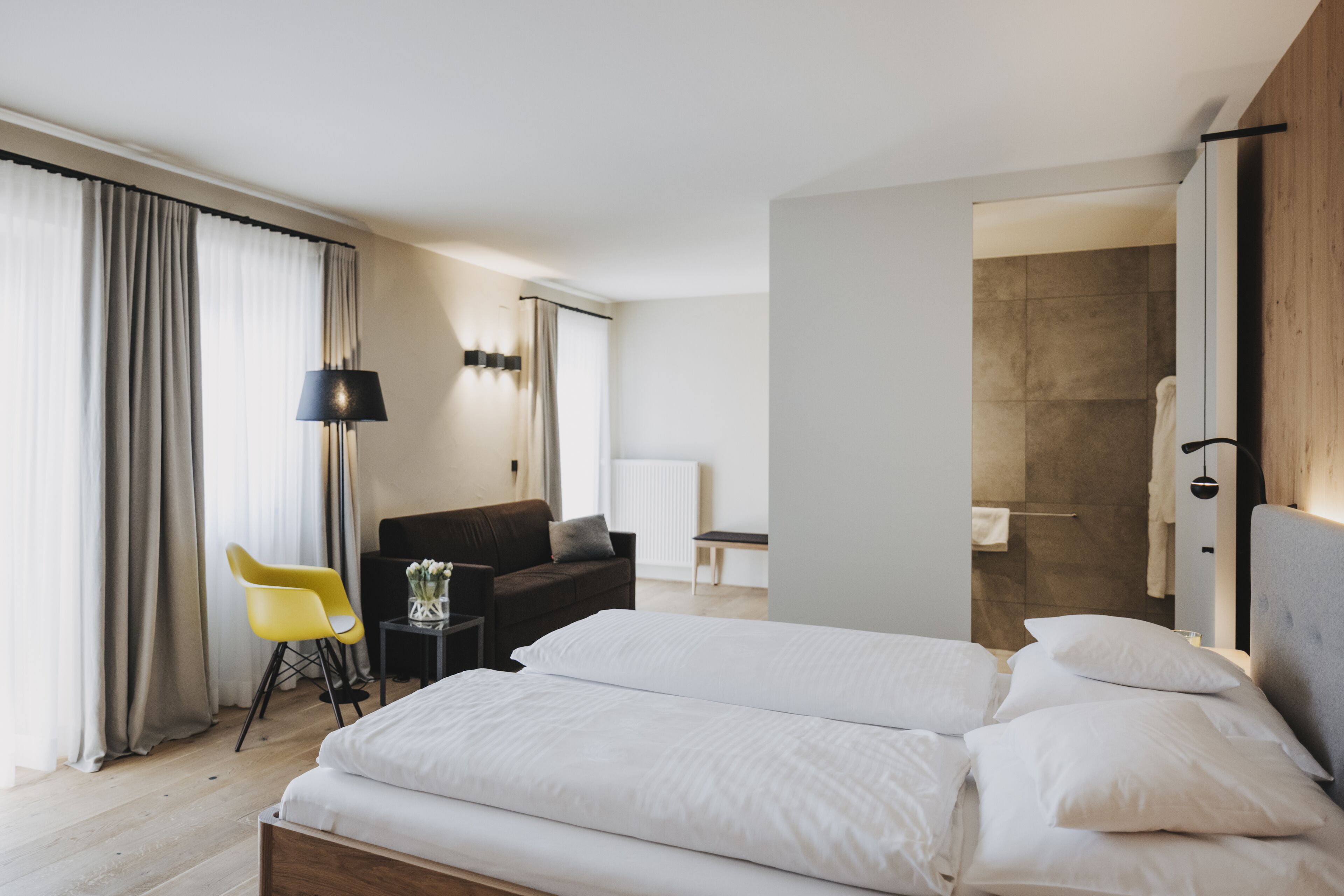A modern hotel room with a large bed, a small desk, a chair, and a shower area.