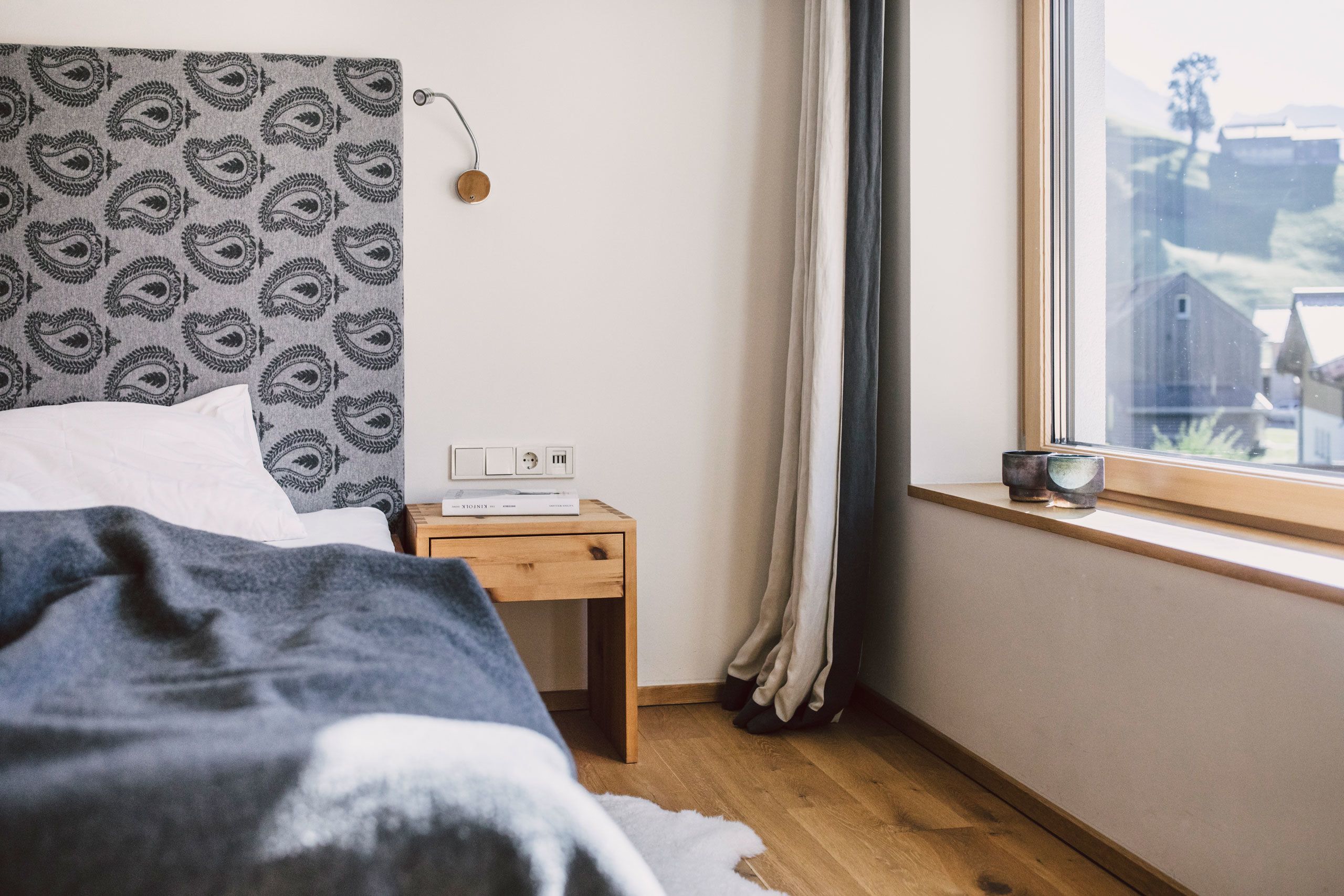 A bedroom with a patterned headboard, a bedside table with a lamp, a window with curtains, and a view of the outside.