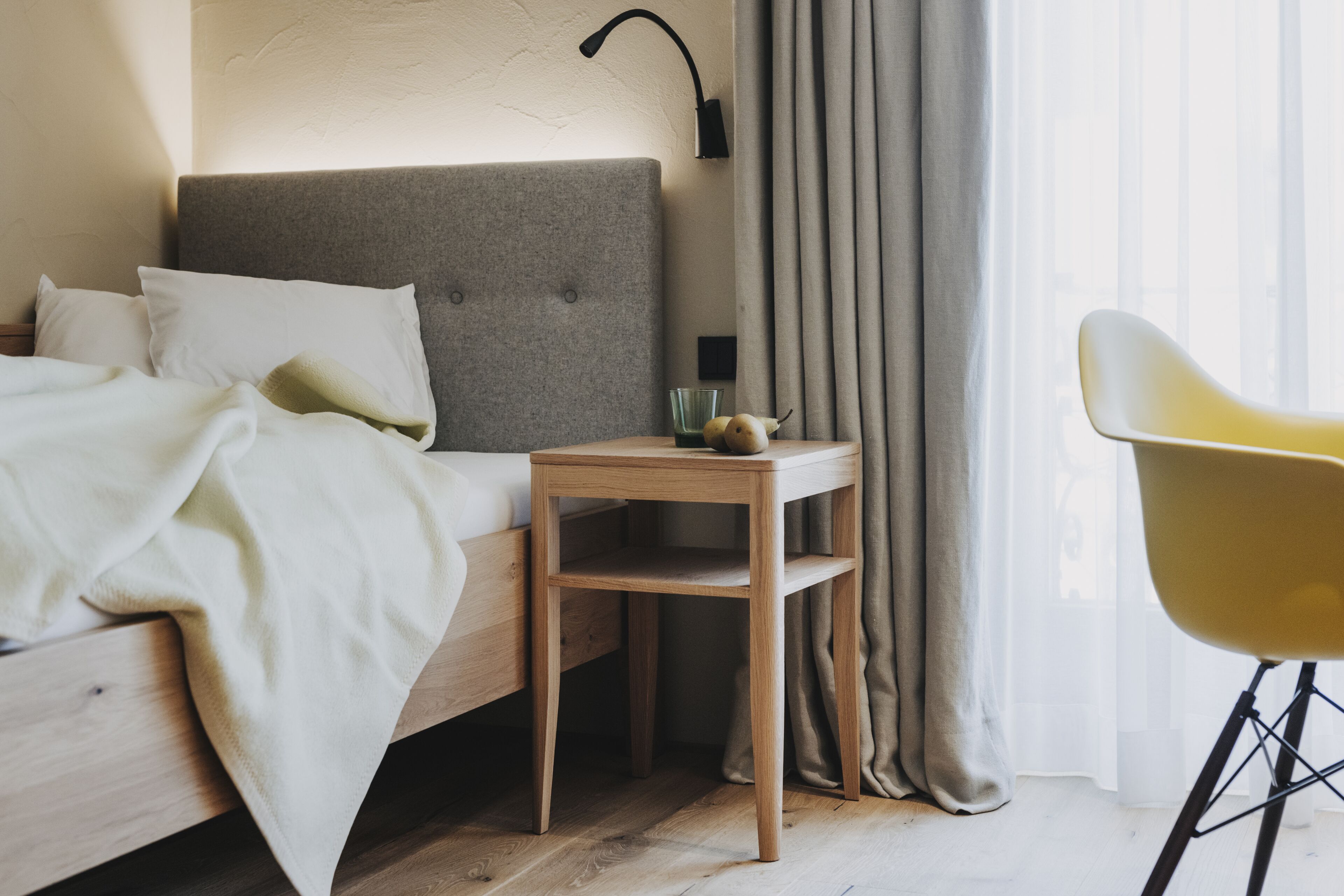 Ein ordentlich bezogenes Bett mit grauem Kopfteil, ein kleiner Nachttisch daneben und ein gelber Sessel in einem hellen Raum mit großem Fenster und transparenten Vorhängen.