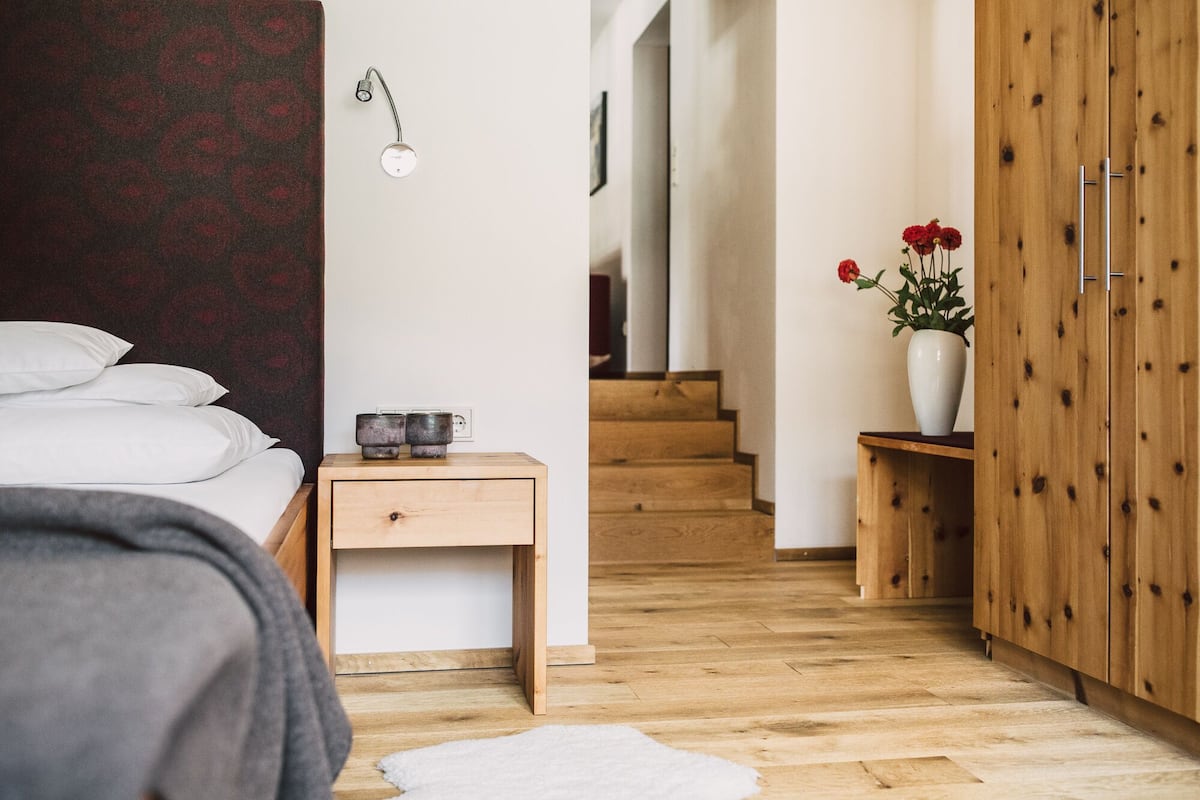 A bedroom with a bed, bedside table, nightstand, wooden wardrobe, and a staircase leading to another room.