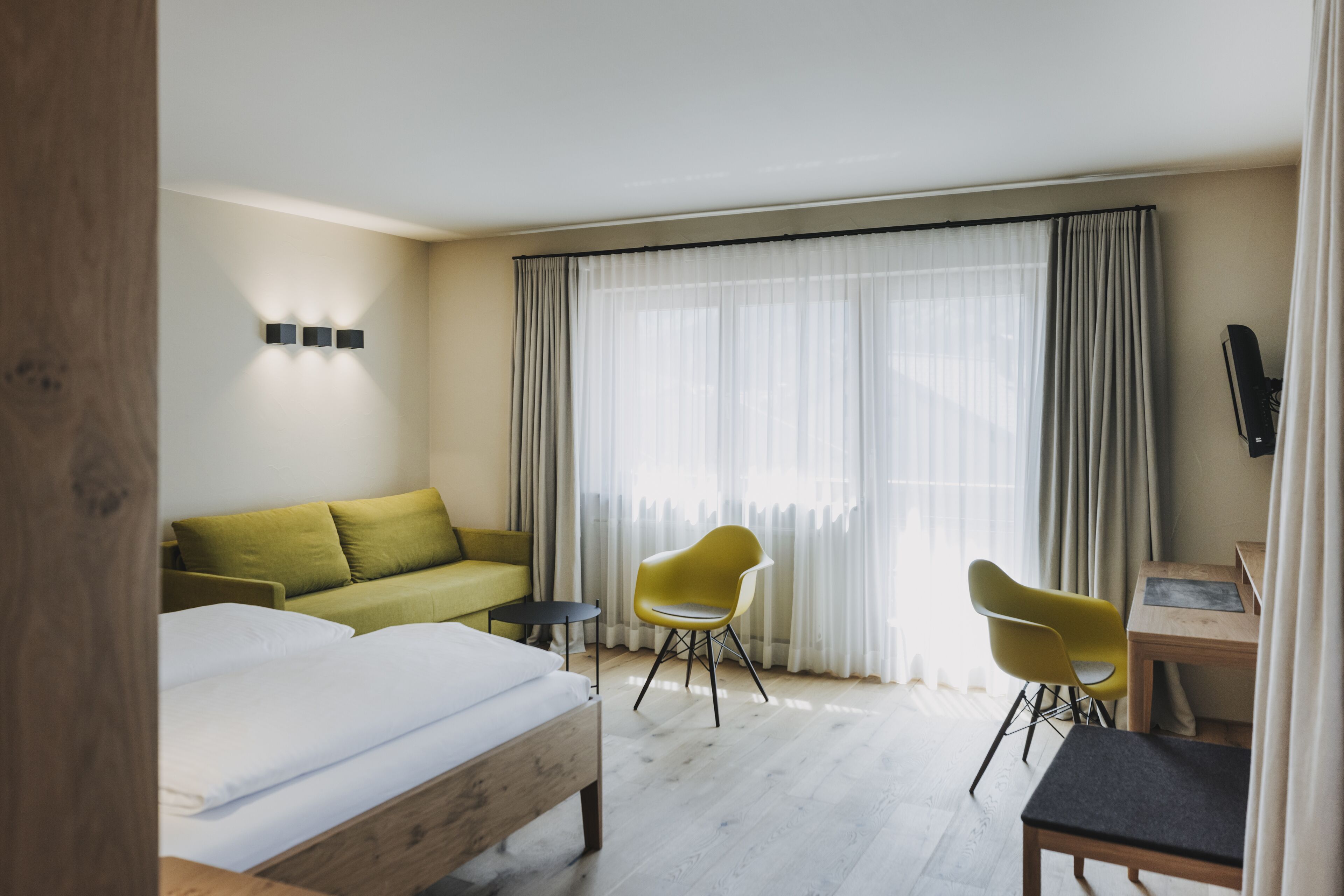 A hotel room with a bed, a green sofa, two yellow chairs, a small table, and a TV.