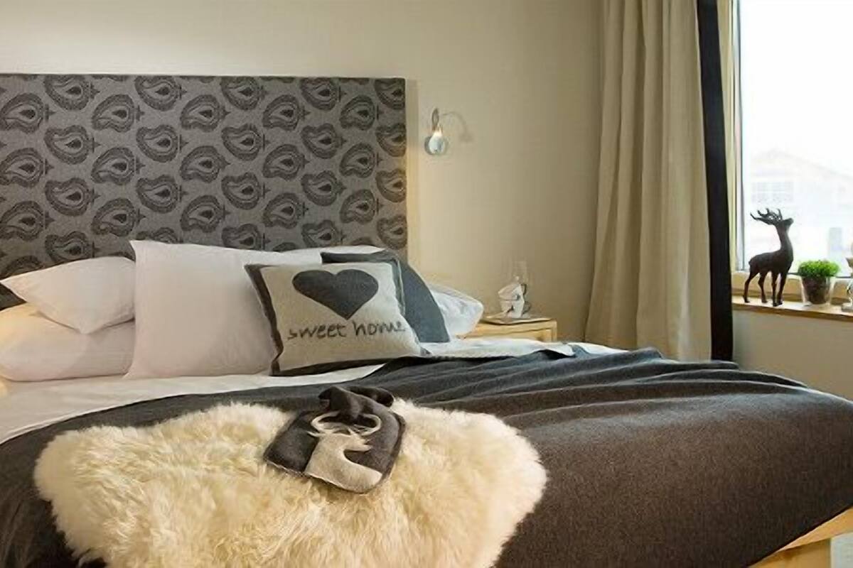 A bedroom with a patterned headboard, a bed with a heart-shaped pillow, and a view of a cityscape through the window.