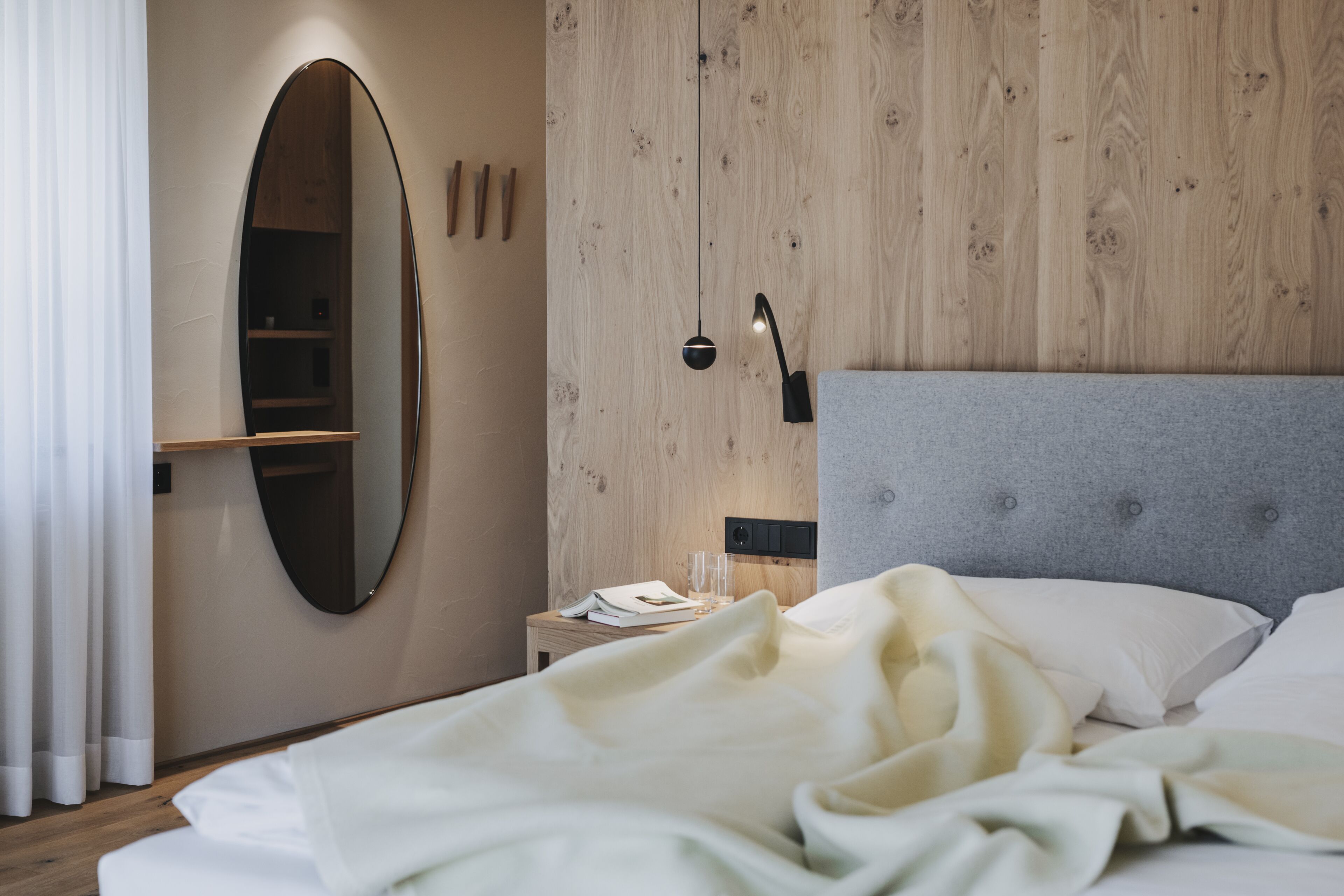 A neatly made bed with white linens, a bedside table with a book and glasses, a mirror, and a wall with wooden paneling.