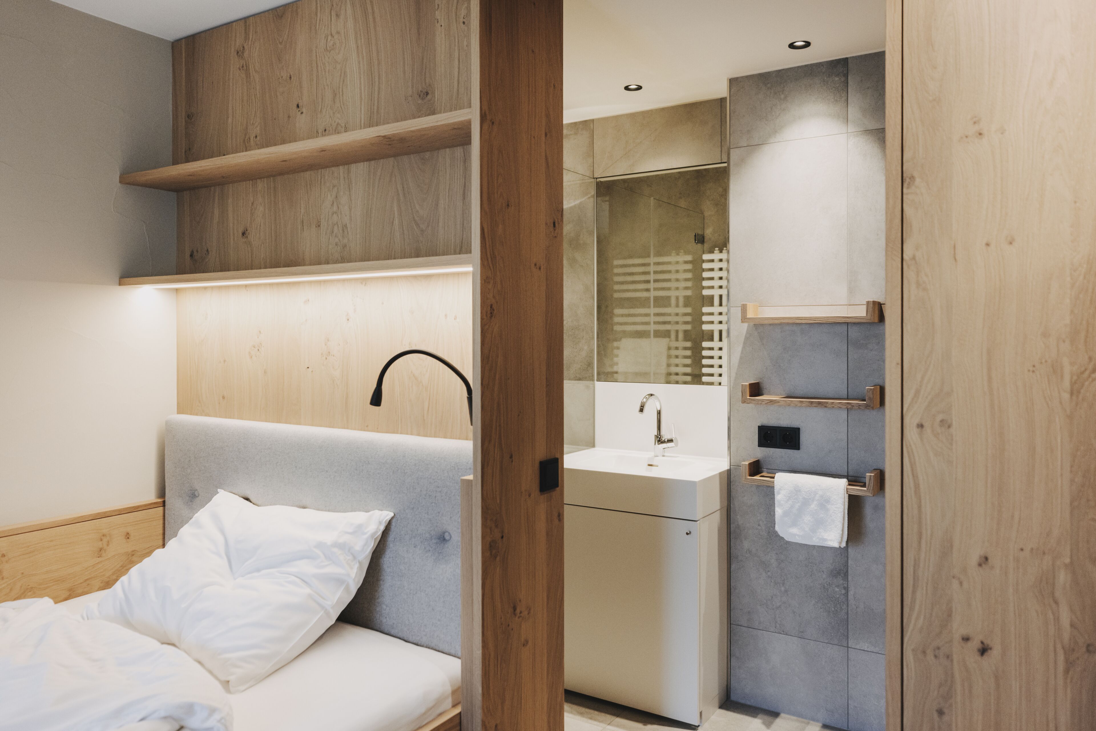 Un dormitorio moderno con cama, cabecera de madera, estante empotrado, mesita de noche y baño con lavamanos y espejo.