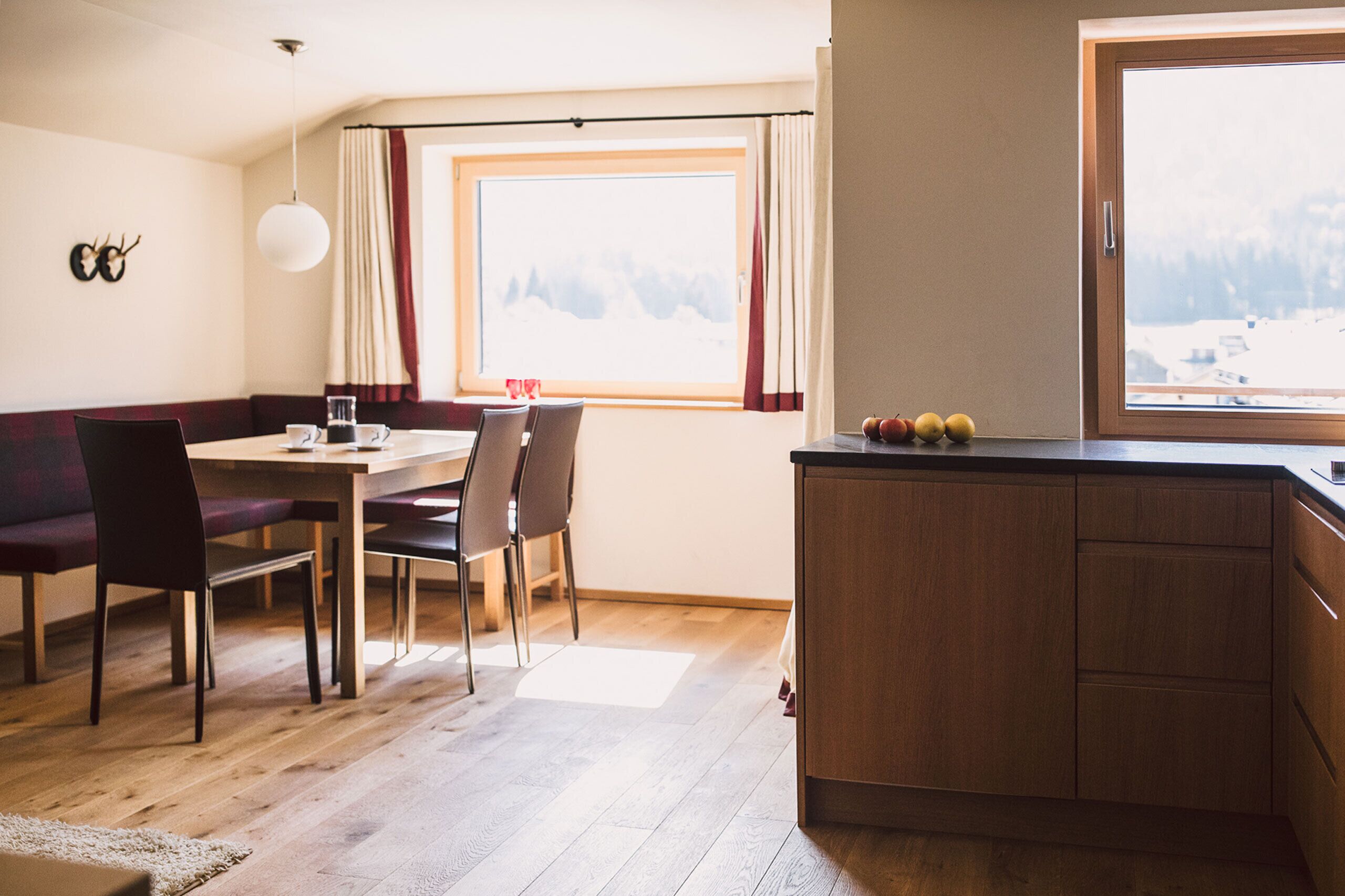Un comedor con una mesa y sillas de madera, un banco, un ventanal con vistas a montañas nevadas y una zona de cocina con un armario de madera y fruta en la encimera.