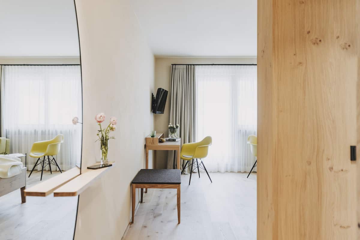 A modern hotel room with a wooden bench, two yellow chairs, a small table with a vase of flowers, and a TV mounted on the wall.