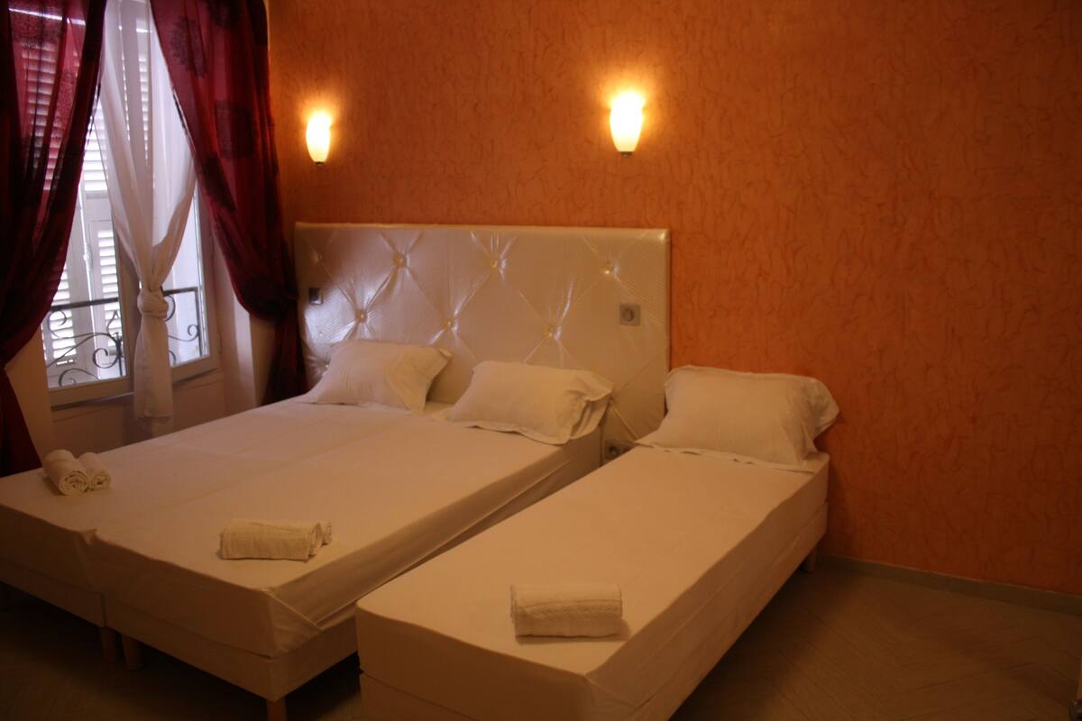 Una habitación de hotel con dos camas individuales, un ventanal con cortinas y lámparas de pared.