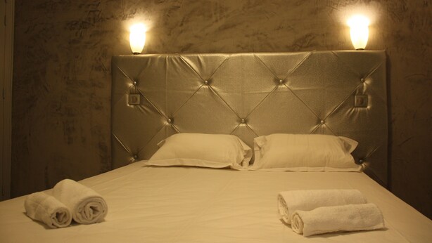 A neatly made bed with two pillows and two rolled towels, a tufted headboard, and wall sconces. - Arcotel (Cannes)