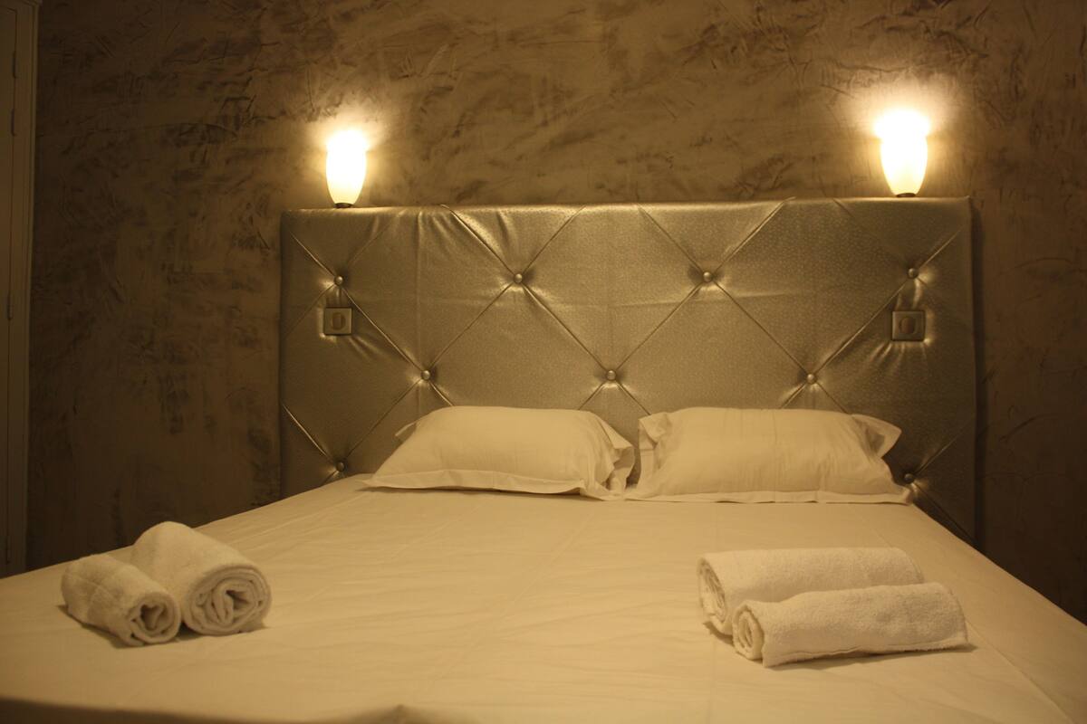 Una cama bien hecha con dos almohadas y dos toallas enrolladas, un cabecero capitonado y apliques de pared.