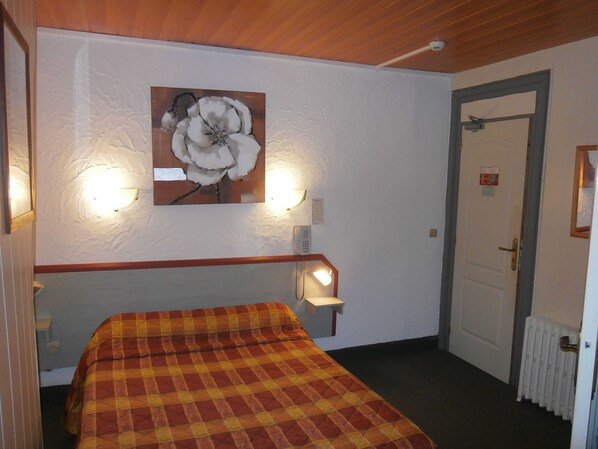 A room with a bed, a wall-mounted lamp, a painting of a flower, and a door. - Hôtel Beauséjour (Rouen)