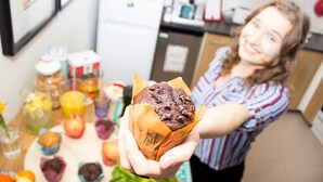 Una persona sostiene un muffin en una cocina con un refrigerador, una cafetera y varios alimentos sobre la encimera.
