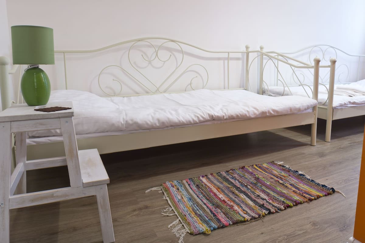 Una habitación con una cama individual de estructura metálica blanca, una mesita de madera con una lámpara verde y una alfombra tejida de colores sobre el suelo de madera.
