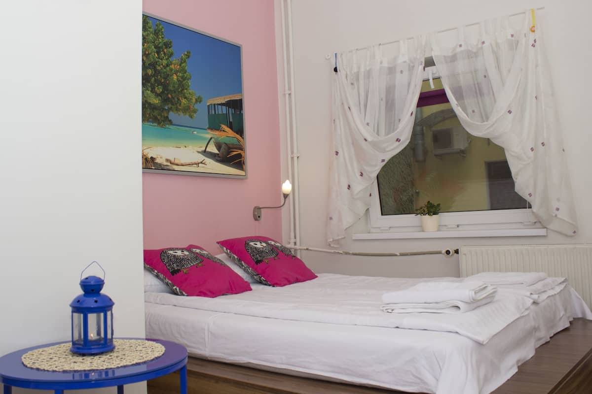 Un dormitorio con una cama, una ventana con cortinas translúcidas, un cuadro de una escena de playa y un farol azul sobre una mesita.