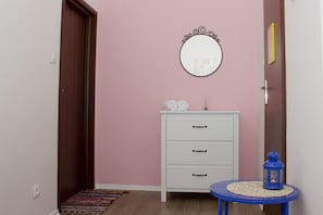 Una habitación con una pared rosa, un aparador blanco de tres cajones, un espejo redondo y un farol azul sobre una mesita.